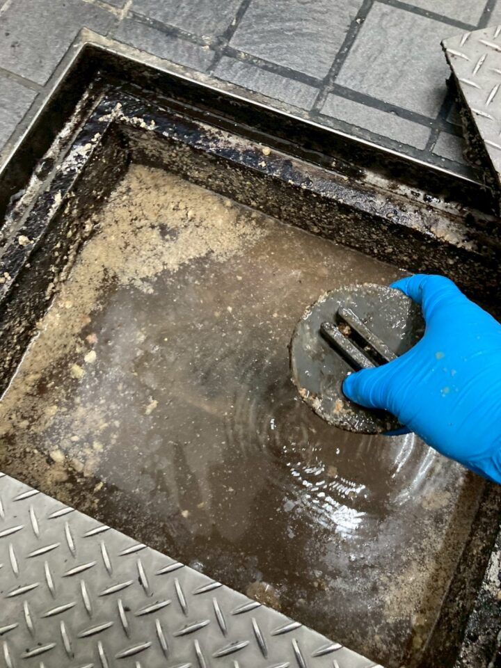 グリストラップ内部の排水管接続キャップを取り外しているところ。青い手袋をした作業者が油脂に覆われた内部で作業