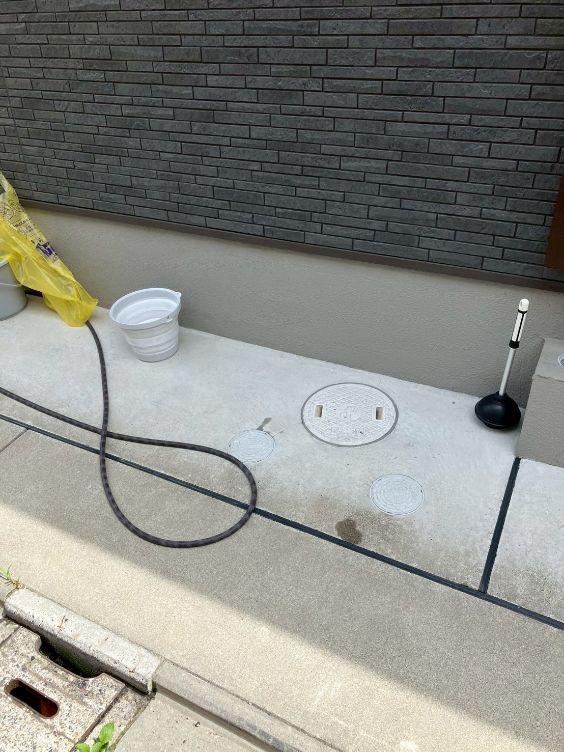 外構コンクリート面に3か所の排水桝蓋が見える。高圧洗浄用の黒ホース・バケツ・ラバーカップが作業準備として並んでいる