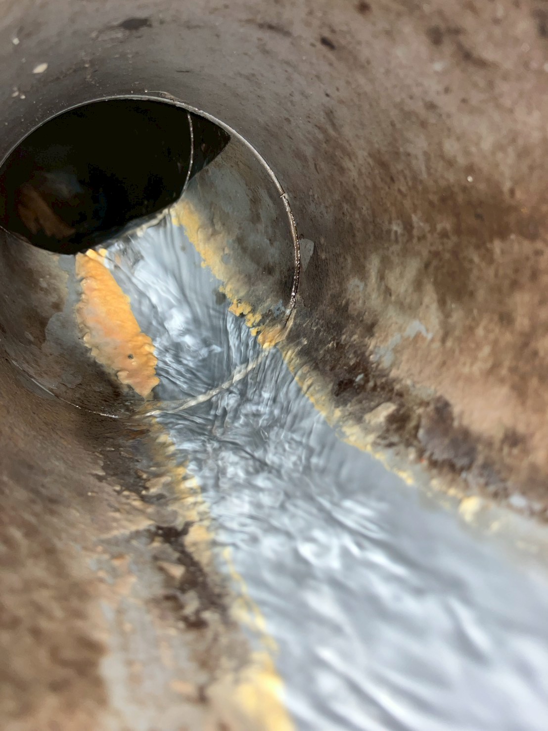 排水桝内部の拡大。内壁は茶褐色のスケールで汚れており、配管接続口付近に黄色〜オレンジ色の油脂固形物が詰まっている。高圧洗浄水が流れ始めている