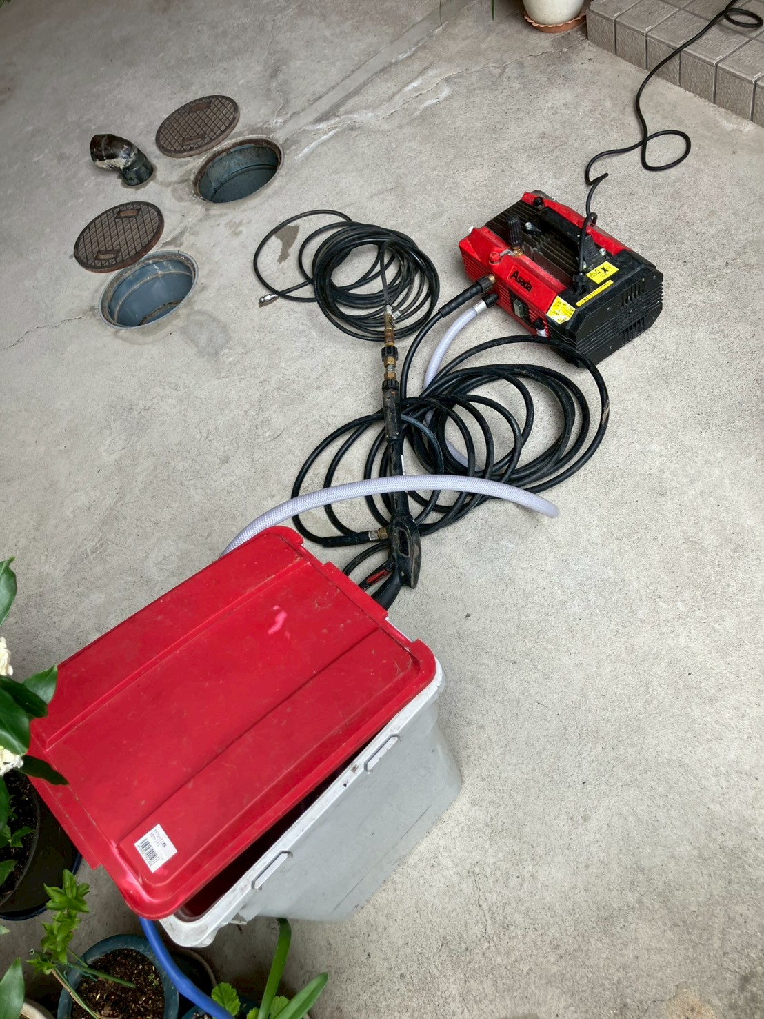 屋外に設置した高圧洗浄機と洗浄ホース。排水桝の蓋が外されており、作業準備が完了した状態