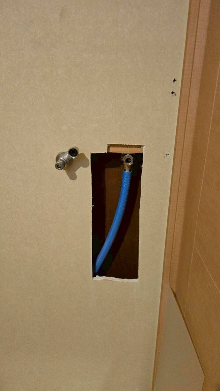 洗濯機置き場の壁を開口し、床下から引き回した青いポリ管を壁内へ引き込んでいる施工中の様子