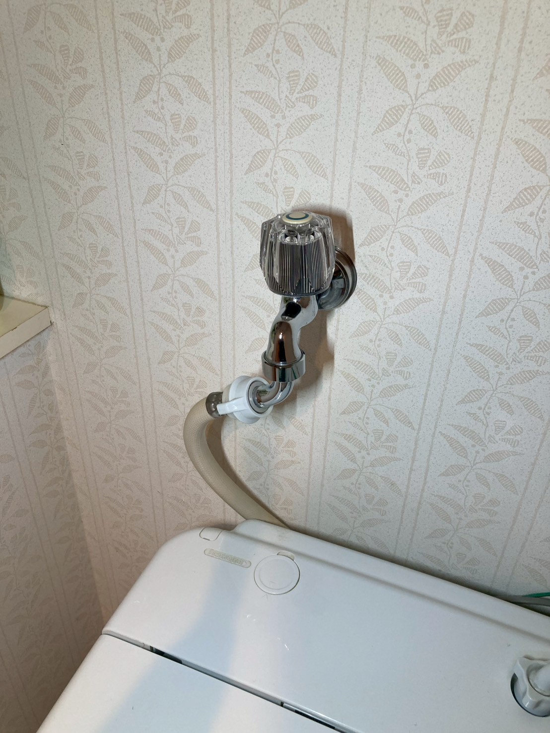 古い透明クリスタルハンドル型の洗濯水栓の全体。洗濯機への給水ホースが接続されている施工前の状態