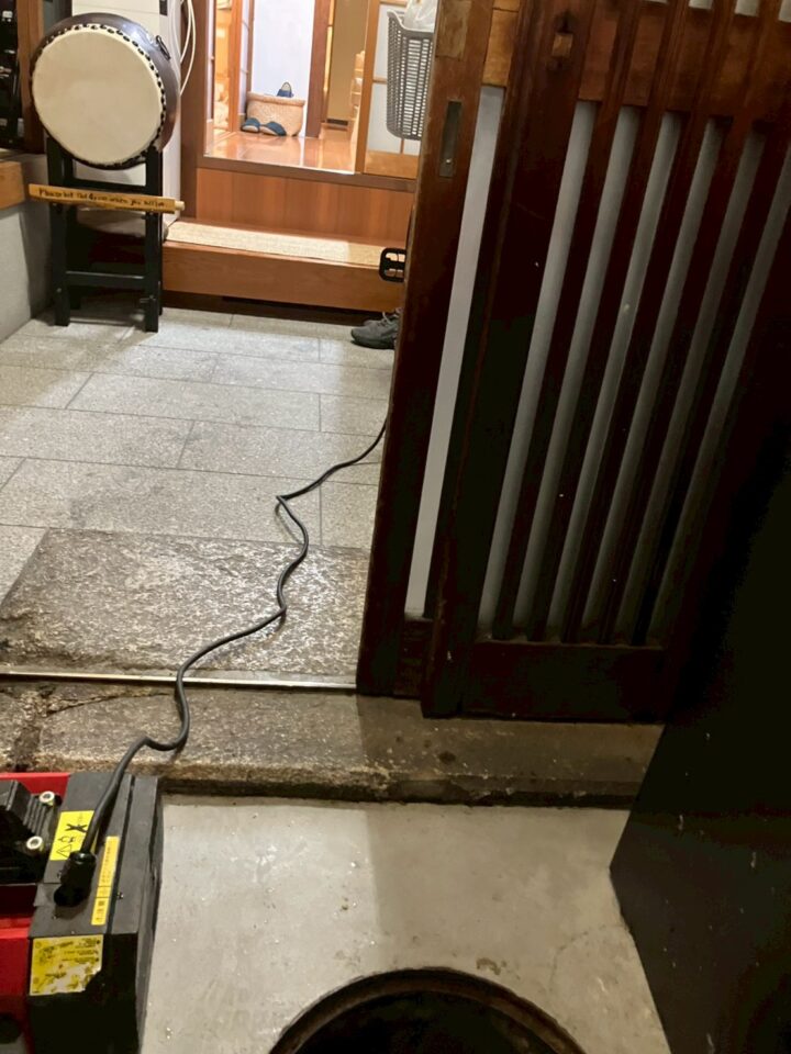 屋内廊下を通じて高圧洗浄ホースが屋外排水枡へ引き込まれている作業中の様子